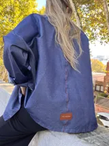 Sobre camisa de gabardina símil denim, color azul, con costuras visibles en color rojo y etiqueta de cuero en la espalda. Presenta un corte holgado y mangas largas.
