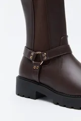 Bota tipo biker de caña media en cuero sintético negro, con panel elástico lateral, suela gruesa dentada y arnés decorativo con herraje metálico.