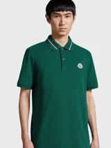 Remera polo Moncler de algodón color verde oscuro, con cuello y puños acanalados con doble raya blanca y roja. Presenta cierre de dos botones y el logo de la marca bordado en el pecho.