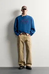 Sudadera azul con corte relaxed fit, cuello redondo y manga larga, confeccionada en tejido de algodón. Presenta un bordado frontal a contraste en color rojo con diseño ornamental y texto.
