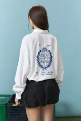 Camisa blanca de corte oversize con cuello camisero y mangas largas. Presenta un bordado en la espalda con el diseño de un escudo nacional estilizado en color azul y la frase 'El país de los mates y las siestas'.