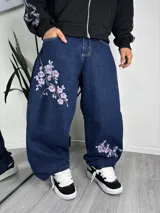 Jeans baggy de denim azul oscuro con diseño bordado de ramas con flores rosadas en una pierna y en el tobillo opuesto.