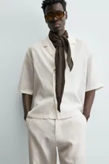 Camisa de lino color blanco, con cuello de solapas de muesca, manga corta y cierre frontal cruzado oculto. Presenta un calce relajado y aberturas laterales en el bajo.