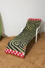 Pareo rectangular de algodón con estampado abstracto en negro y verde sobre fondo blanco. Tiene flecos en los extremos y un borde a cuadros rojos y rosas.