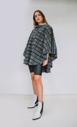 Poncho de paño con estampa a cuadros en tonos grises, negros y blancos, con cuello camisero gris y cierre frontal con dos botones.