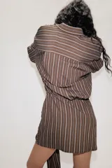 Vestido corto de estilo camisero con estampado de rayas verticales, cuello solapa, manga larga y cierre frontal con botones. Presenta un detalle de lazada en la cintura que crea un efecto drapeado y un bajo asimétrico.