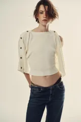 Jersey de punto blanco marfil, con cuello barco y mangas anchas tipo capa decoradas con botones a lo largo de los hombros y brazos.