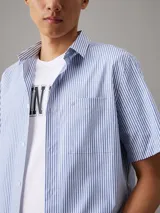 Camisa de manga corta Calvin Klein confeccionada en popelín de algodón elástico con diseño de rayas verticales en blanco y celeste. Presenta cuello en punta, cierre frontal de botones, bolsillo de parche en el pecho y bajo recto.