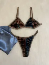Conjunto de lencería compuesto por un bralette de corte triangular y una tanga regulable. Ambas piezas están confeccionadas en terciopelo con un estampado abstracto en tonos azul, marrón y negro, con terminaciones en ribete color marrón.