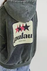 Canguro gris oscuro con efecto lavado (acid wash) y capucha. Presenta un pequeño estampado frontal con estrellas y la palabra "pulau" en tonos rosado y amarillo. Tiene bolsillo canguro y puños acanalados.