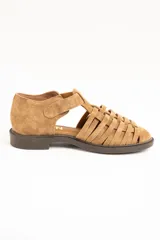 Sandalias cangrejeras de gamuza color visón, con diseño de tiras entrelazadas, tira ajustable con hebilla metálica en el tobillo y suela plana de goma marrón.