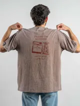 Remera oversize de algodón con efecto lavado, color marrón, con estampado gráfico en la espalda que incluye una cámara fotográfica y la frase 'Freeze the moment'.