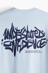 Remera de algodón color celeste con cuello redondo acanalado y hombros caídos. Presenta un estampado gráfico pequeño en el pecho con tipografía estilo graffiti.