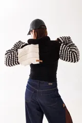 Polera corta de tejido artesanal con diseño bicolor en blanco y negro. Presenta cuello alto, mangas largas con trama a rayas y etiqueta de marca en la parte posterior.
