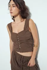 Top corto color marrón, con escote cuadrado y breteles anchos con volados. Presenta frunces horizontales en el torso y una hilera de botones nacarados en el centro frontal.