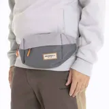 Riñonera Merrell de diseño compacto en dos tonos de gris, con cierre frontal y tiradores en color naranja vibrante. Presenta un parche con el logo de la marca y el año de fundación sobre una base de cuero sintético.