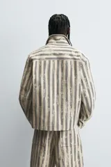 Cazadora de hombre de corte relajado, color beige claro con rayas verticales en azul oscuro con efecto desgastado o estampado. Presenta cuello solapa, manga larga, bolsillos de vivo en cadera y cierre frontal con botones a presión.