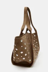 Bolso tipo shopper de hombro, confeccionado en gamuza color marrón, decorado con tachas metálicas plateadas en toda su superficie. Cuenta con doble asa de hombro y cierre mediante botón imantado.
