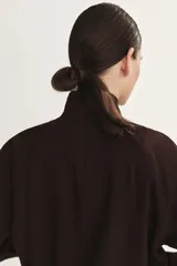 Campera corta oversize de color marrón, confeccionada en tejido de lino. Presenta cuello elevado, cierre frontal oculto con solapa y botones visibles, mangas raglán con puños abotonados, bolsillos delanteros con solapa y cordón ajustable en el bajo.