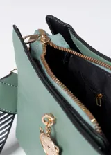 Bandolera verde agua con textura geométrica, cierre superior y correa de tela estampada en blanco y negro. Incluye charm colgante con forma de gato.