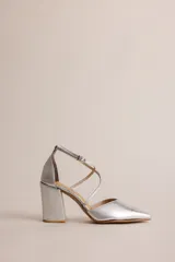 Zapato de tacón color plateado, con punta en V y tiras cruzadas en el empeine que se ajustan al tobillo. El tacón es grueso y cuadrado.