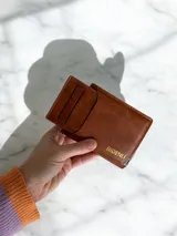 Billetera de eco cuero color marrón con tarjetero extraíble integrado. Presenta un diseño compacto con ranuras para tarjetas y logo grabado en tono dorado.