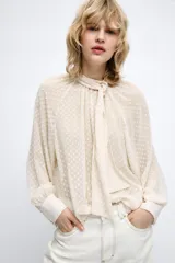 Camisa cruda de manga larga con textura plumeti, cuello redondo y lazada en el mismo tejido. Cuenta con mangas abullonadas terminadas en puños anchos y cierre en la espalda con abertura y botones.