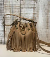Cartera tipo bucket bag color marrón, confeccionada en cuero con textura granulada. Presenta un diseño decorado con flecos largos en todo el frente, herrajes metálicos en tono bronce envejecido, cierre mediante cordón ajustable y correa larga regulable para llevar al hombro o cruzada.