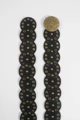Cinturón de cuero sintético negro formado por piezas circulares entrelazadas, decoradas con ojalillos y tachas metálicas doradas. Cuenta con una hebilla circular grande con diseño grabado.