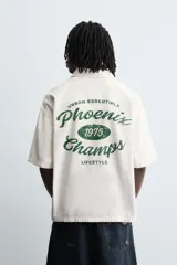 Camisa de algodón con estructura canvas, color rojo lavado, de corte relajado (relaxed fit) y manga corta. Posee cuello solapa, dos bolsillos de plastrón en el pecho y cierre frontal de botonadura. Presenta bordado y estampado de texto a contraste en el delantero.