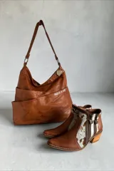Cartera de cuero natural en color marrón, con diseño envolvente, caída suave y bolsillo exterior cruzado con el nombre de la marca bordado. Cuenta con tiras de hombro, herrajes metálicos y un dije colgante geométrico.