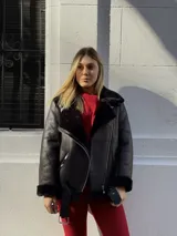 Campera tipo gamulán de cuero sintético negro con interior y detalles de corderito al tono. Presenta cierre frontal diagonal, cuello amplio, bolsillos laterales con cierre y cinturón ajustable en el ruedo.