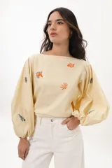 Blusa color arena de algodón con escote a la base y mangas abullonadas. Presenta bordados artesanales de motivos marinos en tonos naranja y azul.