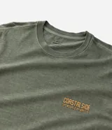 Remera de hombre, modelo comfort, color verde oliva desteñido, con cuello redondo y manga corta. Presenta un pequeño estampado en el pecho con lettering en tonos naranjas que dice "COASTALSIDE".