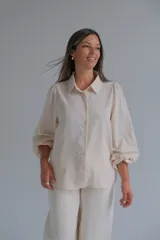 Camisa de color beige con mangas abullonadas y puños ajustados. Presenta cuello clásico, cierre frontal con botones y detalles bordados sutiles en las mangas.