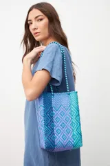 Bolso tipo matera tejido artesanalmente con tiras de plástico en tonos degradados de azul, violeta y turquesa, con diseño geométrico y correas trenzadas.