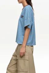 Remera estilo boxy de algodón con diseño de rayas horizontales verdes sobre fondo crudo. Cuenta con cuello redondo marrón tostado, mangas cortas caídas y detalle de logo bordado en el pecho.