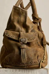 Cartera de ante color marrón con terminación gastada, cuerpo blando, doble asa de mano, cierre frontal con hebilla y detalles de tachas. Incluye cordones de ajuste en los laterales y un colgante metálico decorativo.
