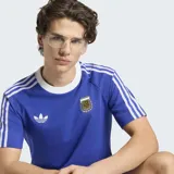 Remera de algodón y poliéster color azul, con cuello redondo blanco, mangas cortas con las tres tiras características de la marca en blanco, logo del trifolio y escudo de la AFA en el frente.