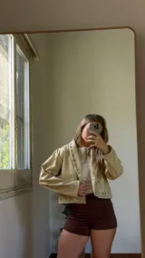 Campera corta de ecocuero color crudo, con diseño de solapas con ojales metálicos decorativos y cierre frontal.