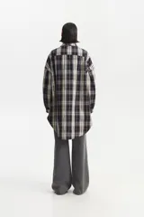 Camisa de largo extendido, estilo camisaco, con patrón de cuadros escoceses en tonos negro, blanco crema y gris lavanda. Presenta cuello camisero, cierre frontal con botones y dos bolsillos de parche con tapa en el pecho. El corte es holgado y la parte inferior es redondeada.
