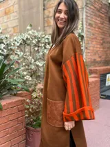 Saco largo de lana merino en color marrón, con diseño abierto sin botones. Presenta mangas anchas con estampado de rayas verticales en tonos naranja y marrón, y un bolsillo frontal aplicado en material símil cuero con textura.