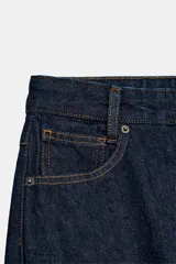 Jeans de tiro bajo y pernera ancha, confeccionados en denim azul oscuro con pespuntes en contraste. Cuentan con un diseño de cinco bolsillos, cierre frontal con cremallera y botón metálico, y destacan por su amplio doblando en el bajo que deja ver el revés de la tela con detalles de costura selvedge.