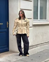 Chaqueta estructurada color beige con cuello, ribetes de bolsillos y detalles en negro. Presenta un diseño entallado con cierre frontal completo y hombreras marcadas.