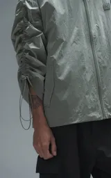 Campera rompevientos de diseño oversize, color verde, confeccionada en microfibra técnica ligera. Presenta capucha integrada, cierre frontal técnico y un sistema de frunces elásticos regulables a lo largo de las mangas que permiten ajustar el volumen de la prenda.