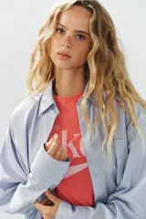 Camisa oversize de manga larga con cuello solapa, cierre frontal de botones y bolsillo de parche en el pecho, con diseño de rayas verticales celestes y blancas.