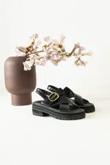 Sandalias negras con diseño de tiras cruzadas en cuero con textura de reptil y detalles en gamuza. Poseen una hebilla metálica dorada ajustable en el lateral y una suela track robusta de goma.