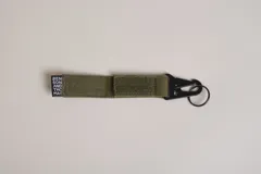 Llavero de cinta de poliéster color verde oliva con mosquetón y aro metálicos negros. Incluye etiqueta con el logo de la marca.