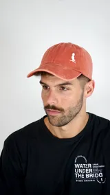 Gorra de béisbol color salmón con efecto desgastado, presenta un pequeño bordado blanco en el frente que representa un pájaro sobre una rama.