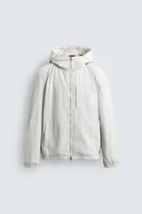Cazadora de ajuste relajado, color blanco roto o gris muy claro, confeccionada en tejido técnico. Presenta cuello subido con capucha, cierre frontal de cremallera, bolsillo de parche en el pecho y bolsillos laterales de vivo. Los puños y el bajo terminan en elástico.